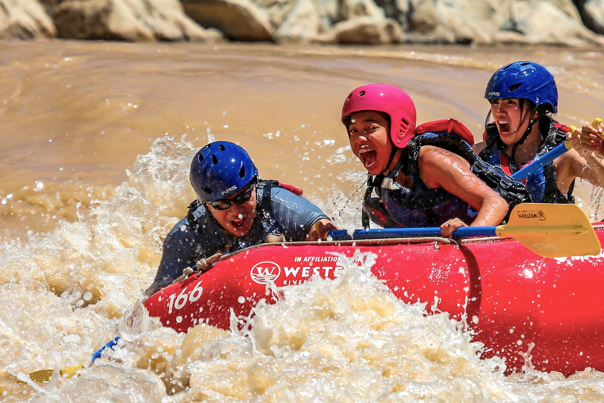 Three people whitewater rafting in a red raft; splashing through rough water, wearing helmets and life vests.