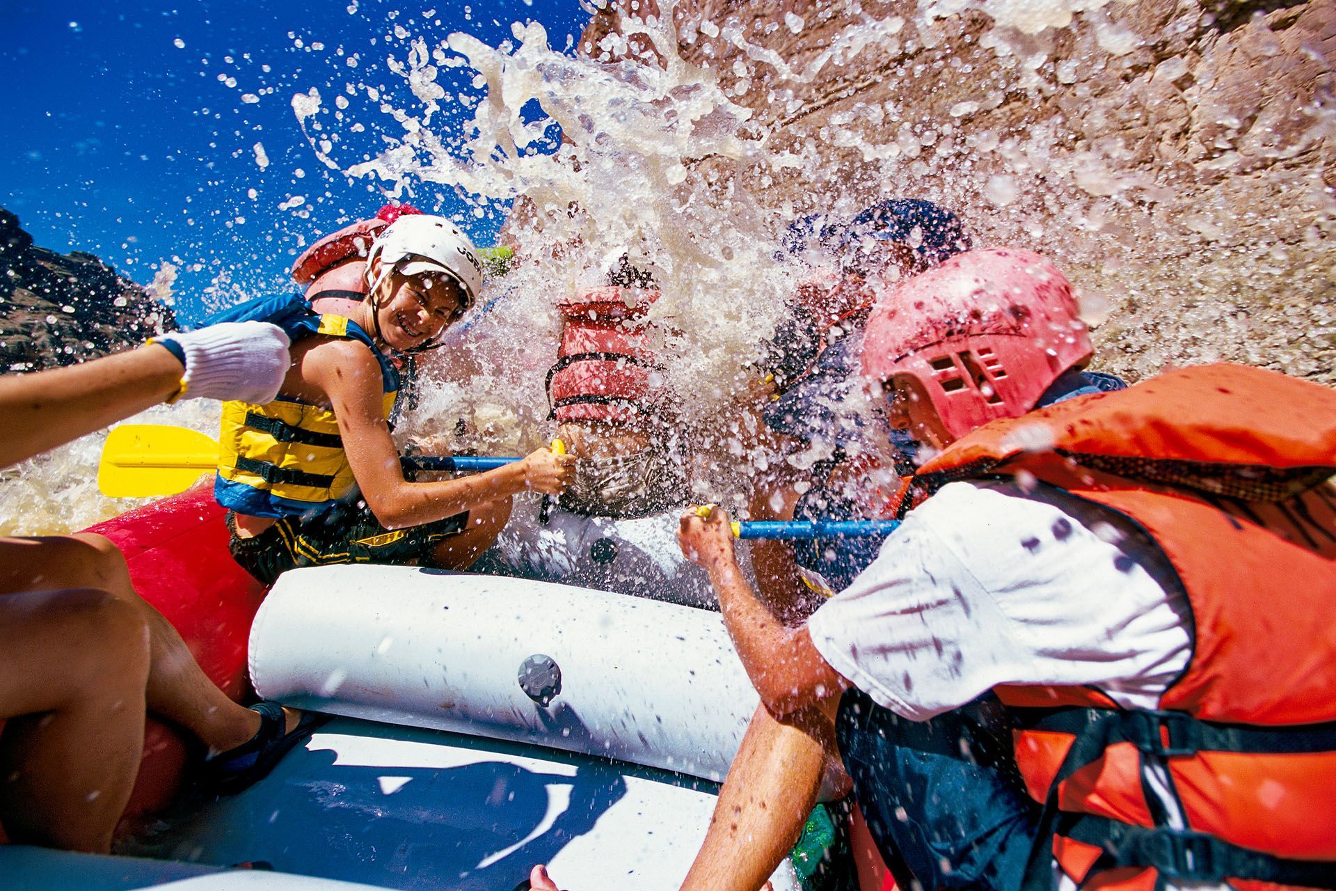 Whitewater rafting with splashing water; people in life vests and helmets, paddling.