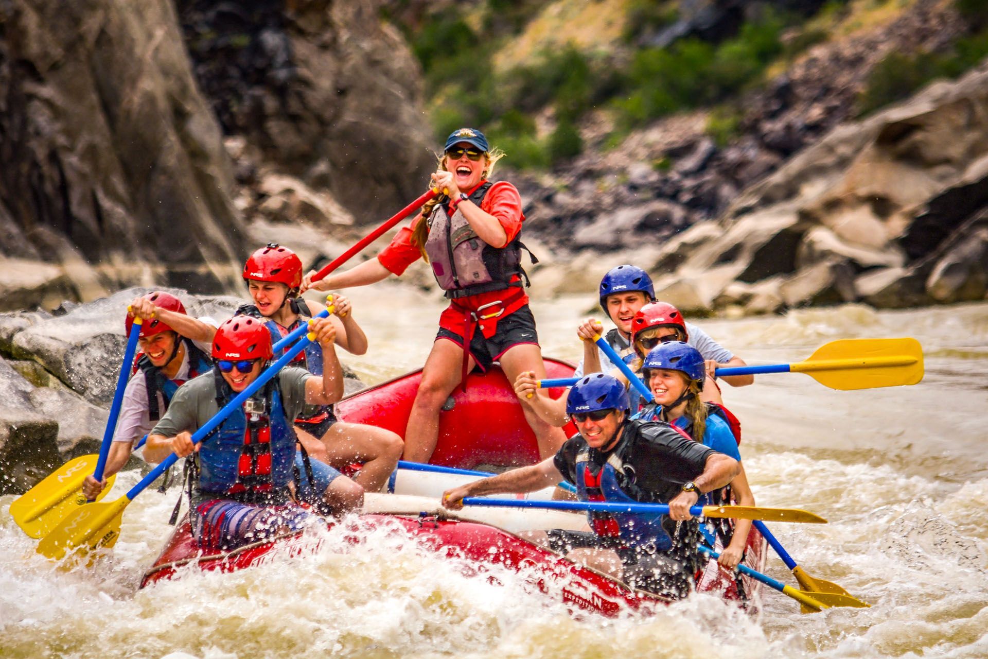 People white water rafting through rapids on Westwater Canyon, holding paddles, wearing life vests and helmets.