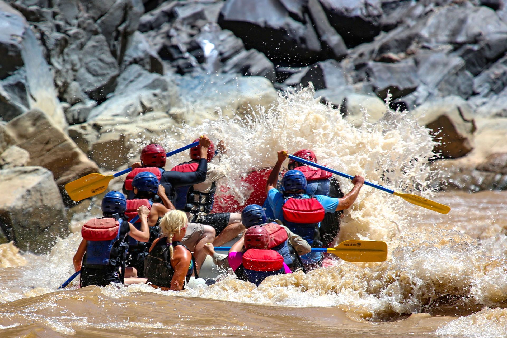 People white water rafting through a large wave, water splashing.