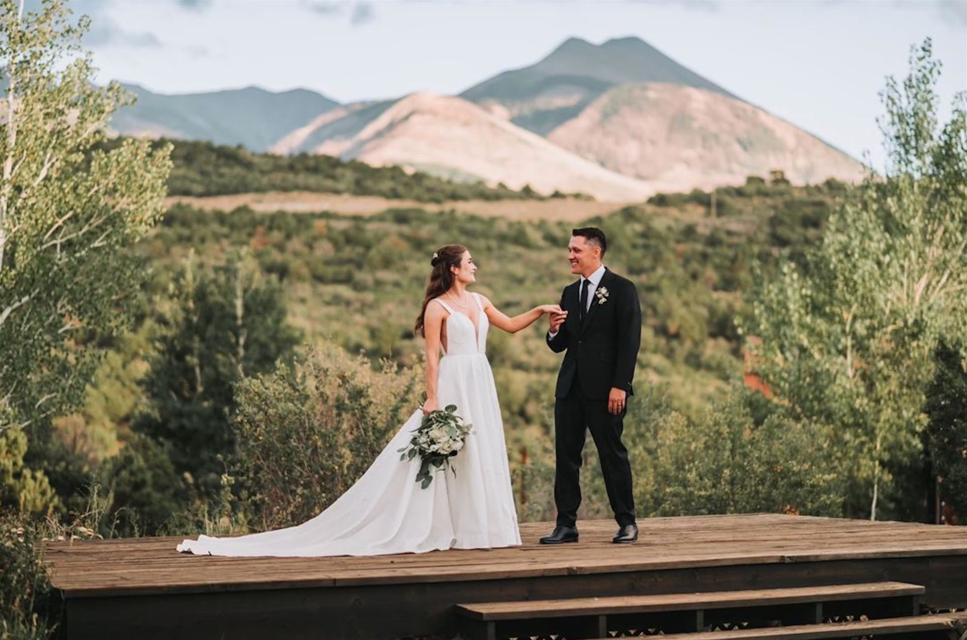 Bride and groom on a wooden platform with mountains in the background; bride holding a bouquet.