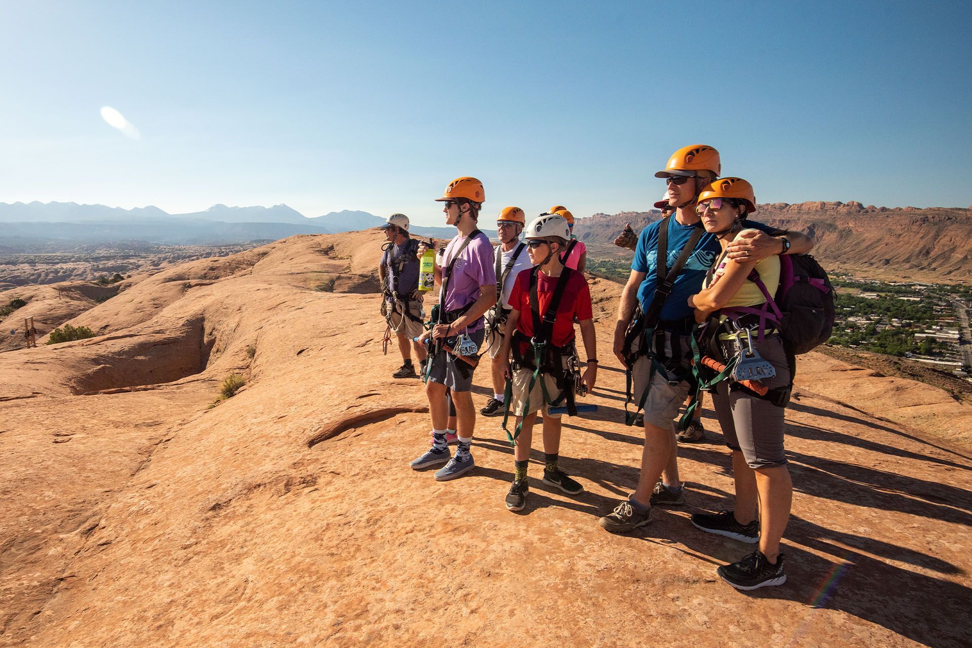 Group of people in helmets and harnesses on a red rock, overlooking a valley under a blue sky.