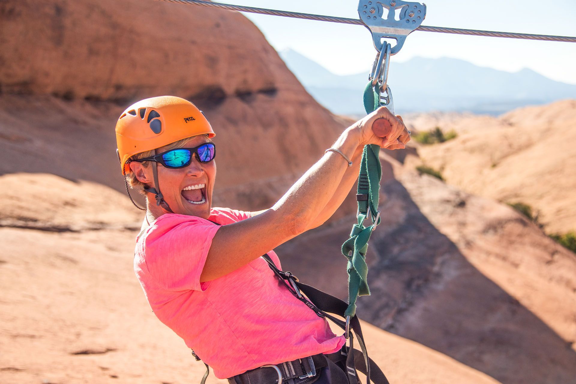 Person zip-lining with an orange helmet, pink shirt, and sunglasses, Moab desert background.