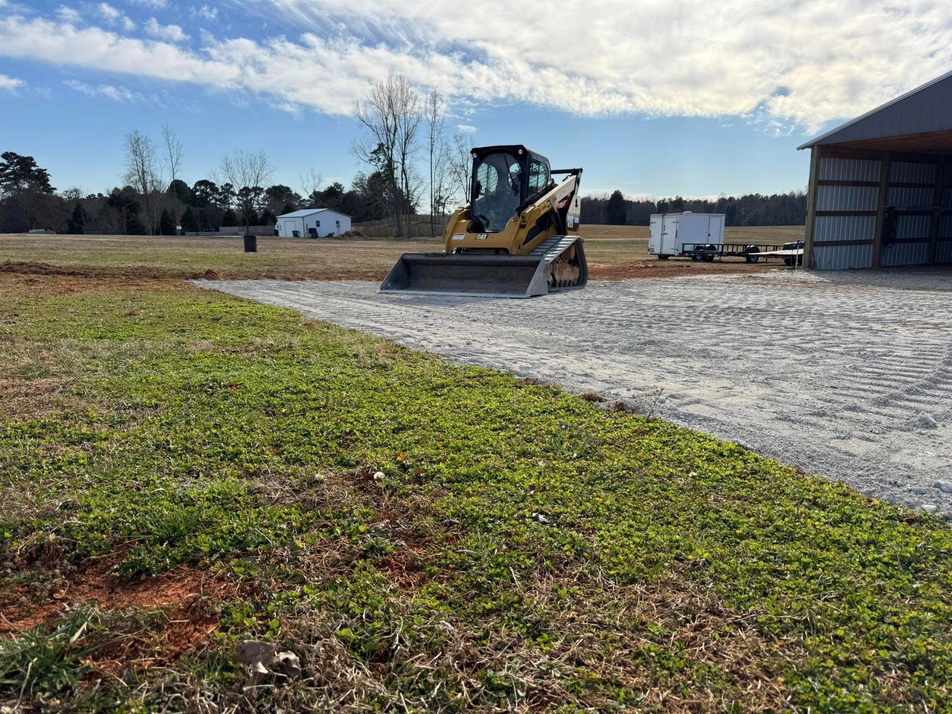 Yellow skid steer on gravel near a shed in a rural field under a cloudy sky