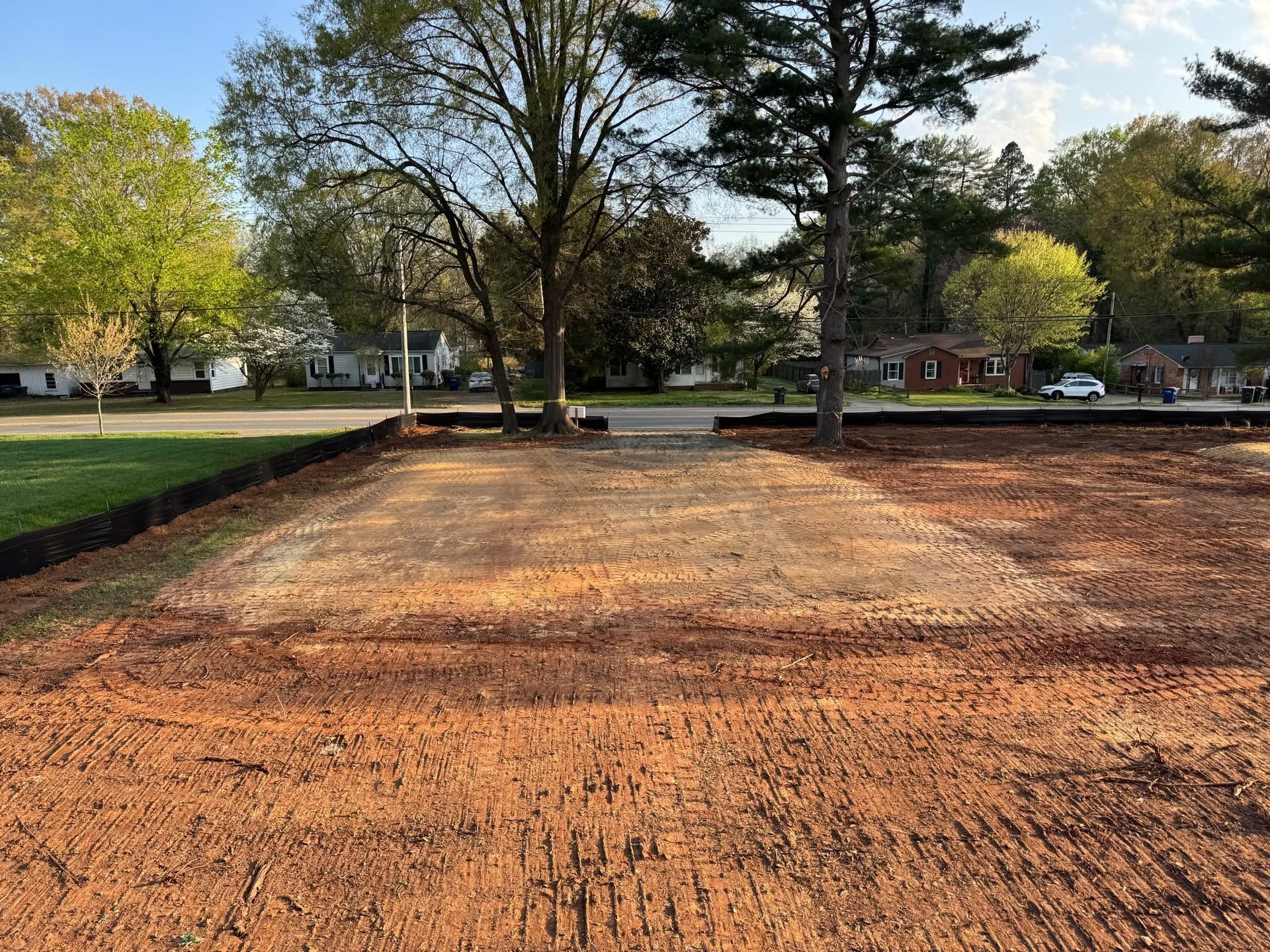 Freshly leveled dirt area in a park, bordered by grass and trees under a clear sky.