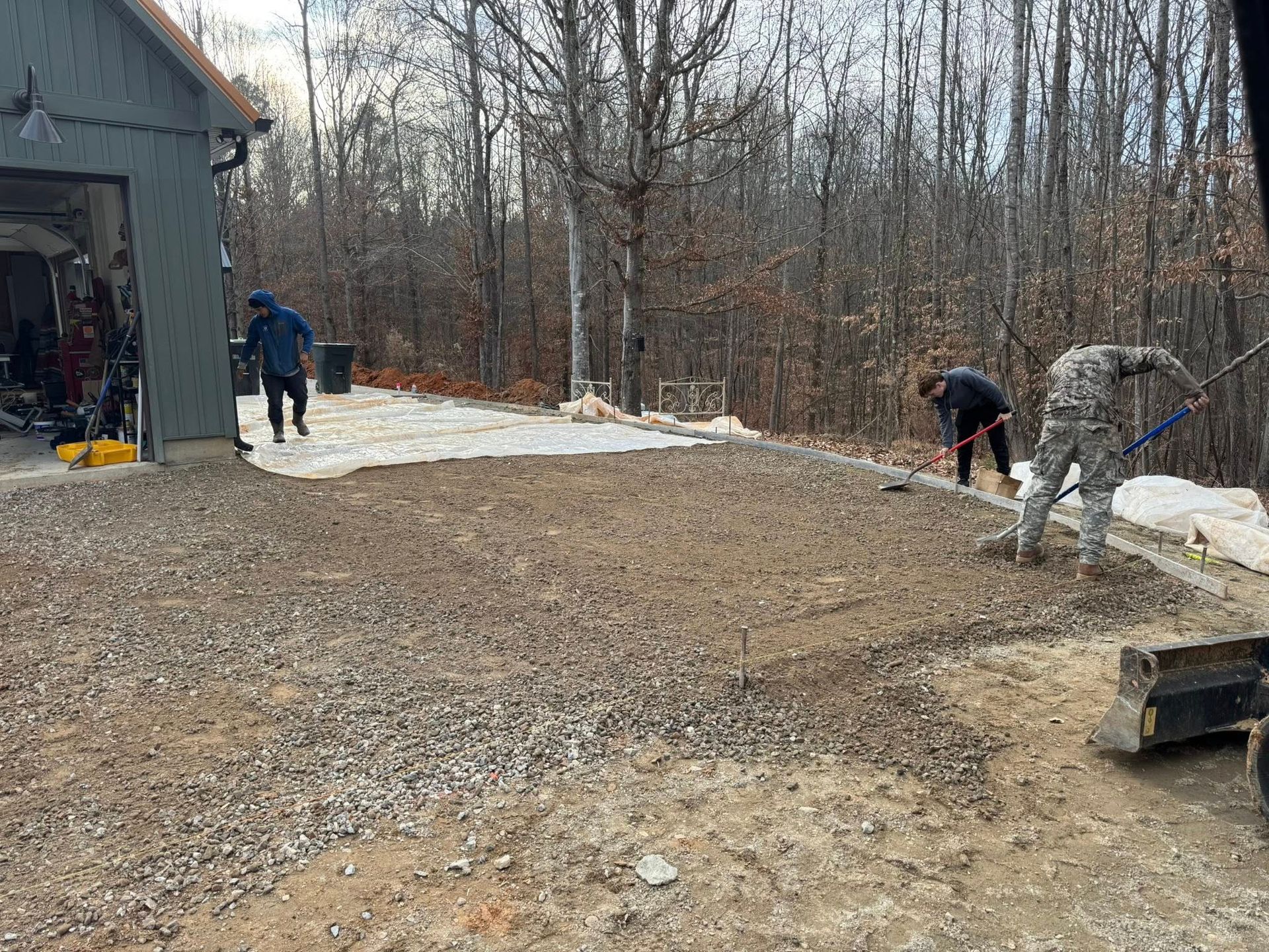 Workers leveling gravel driveway beside a house in a snowy, wooded yard