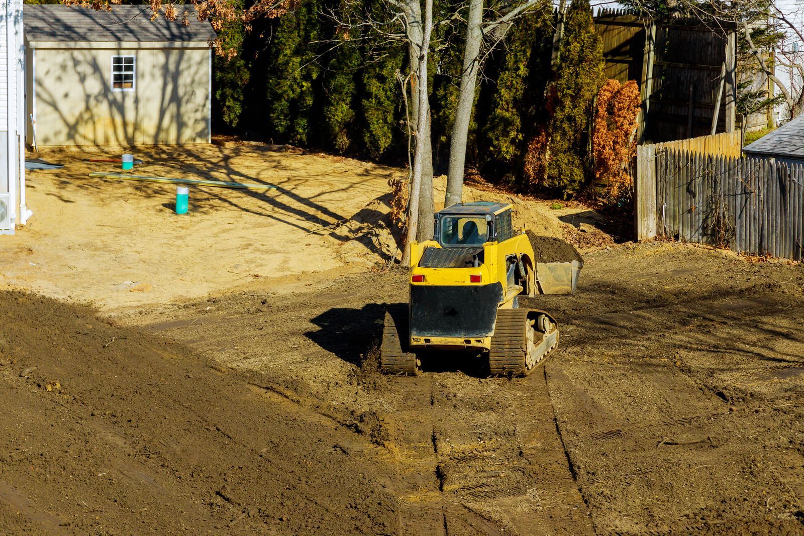 Yellow bulldozer grading a dirt lot near a house and trees, with a fence in the background