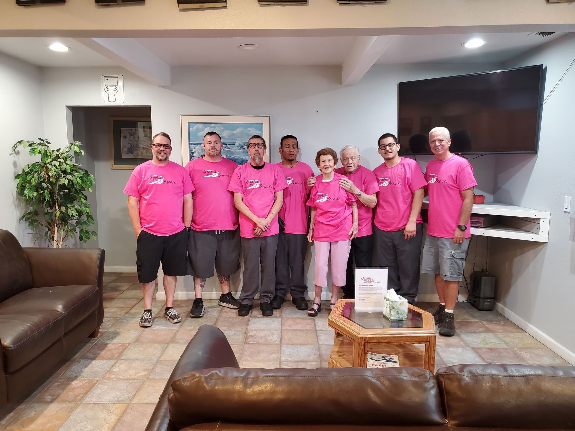 Group of nine people in pink shirts posing indoors. They stand in a living room setting with a couch and TV. | Hal's Auto Care