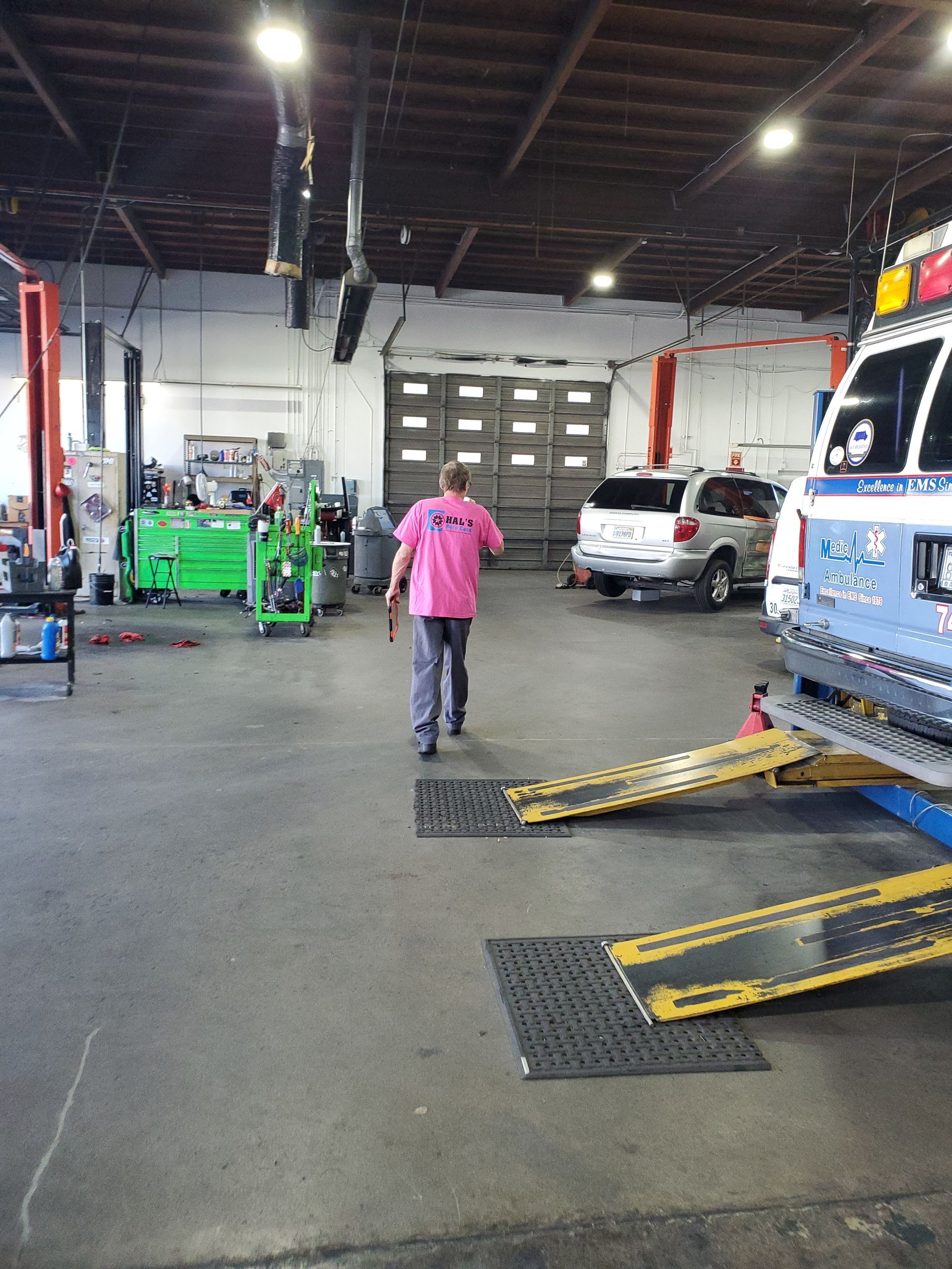 A man in pink shirt walks in a garage. A car is on a lift, and a green toolbox is in the background. | Hal's Auto Care