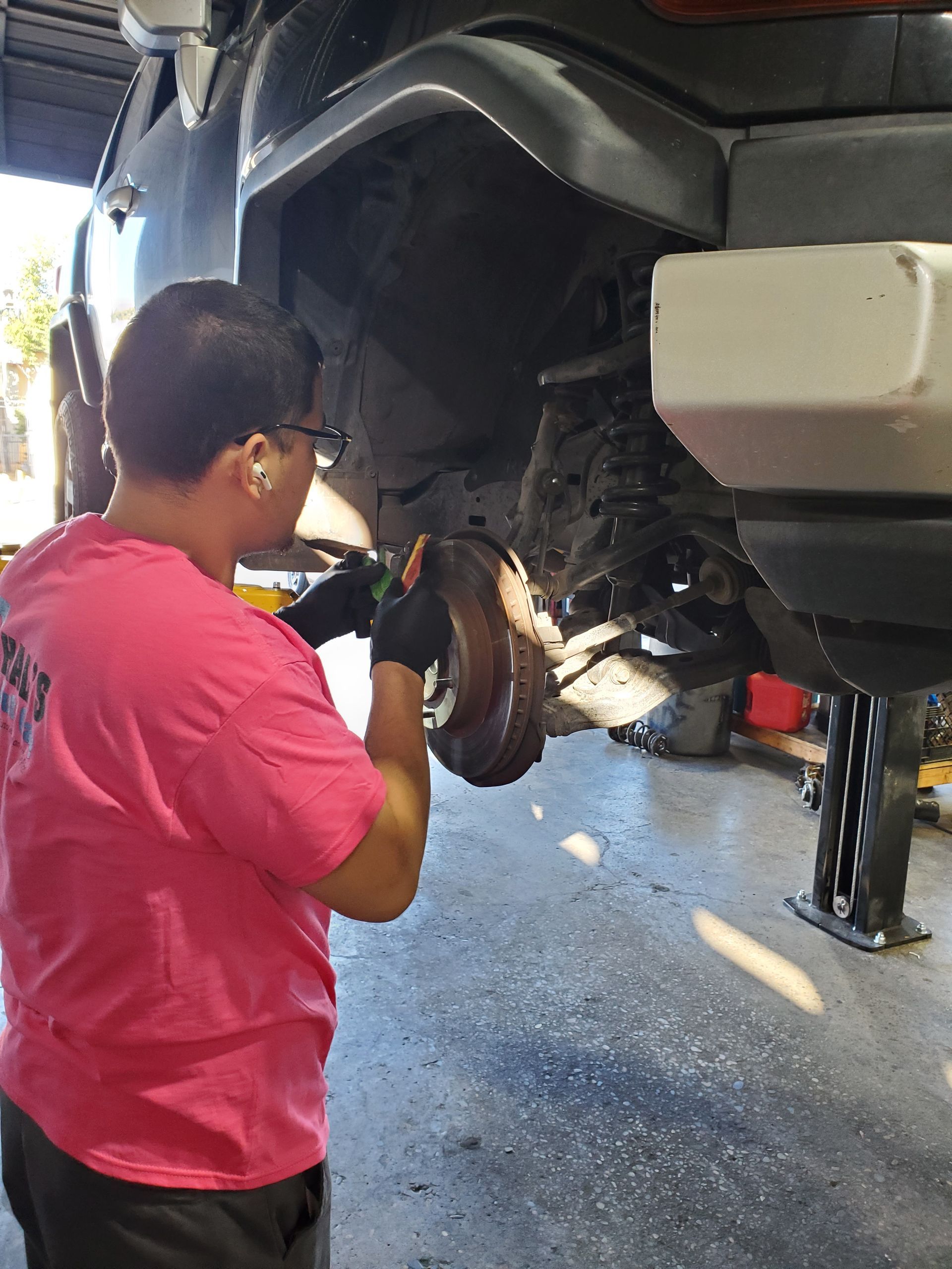 Mechanic in pink shirt holding a rusty brake drum, working under a lifted car in a shop. | Hal's Auto Care