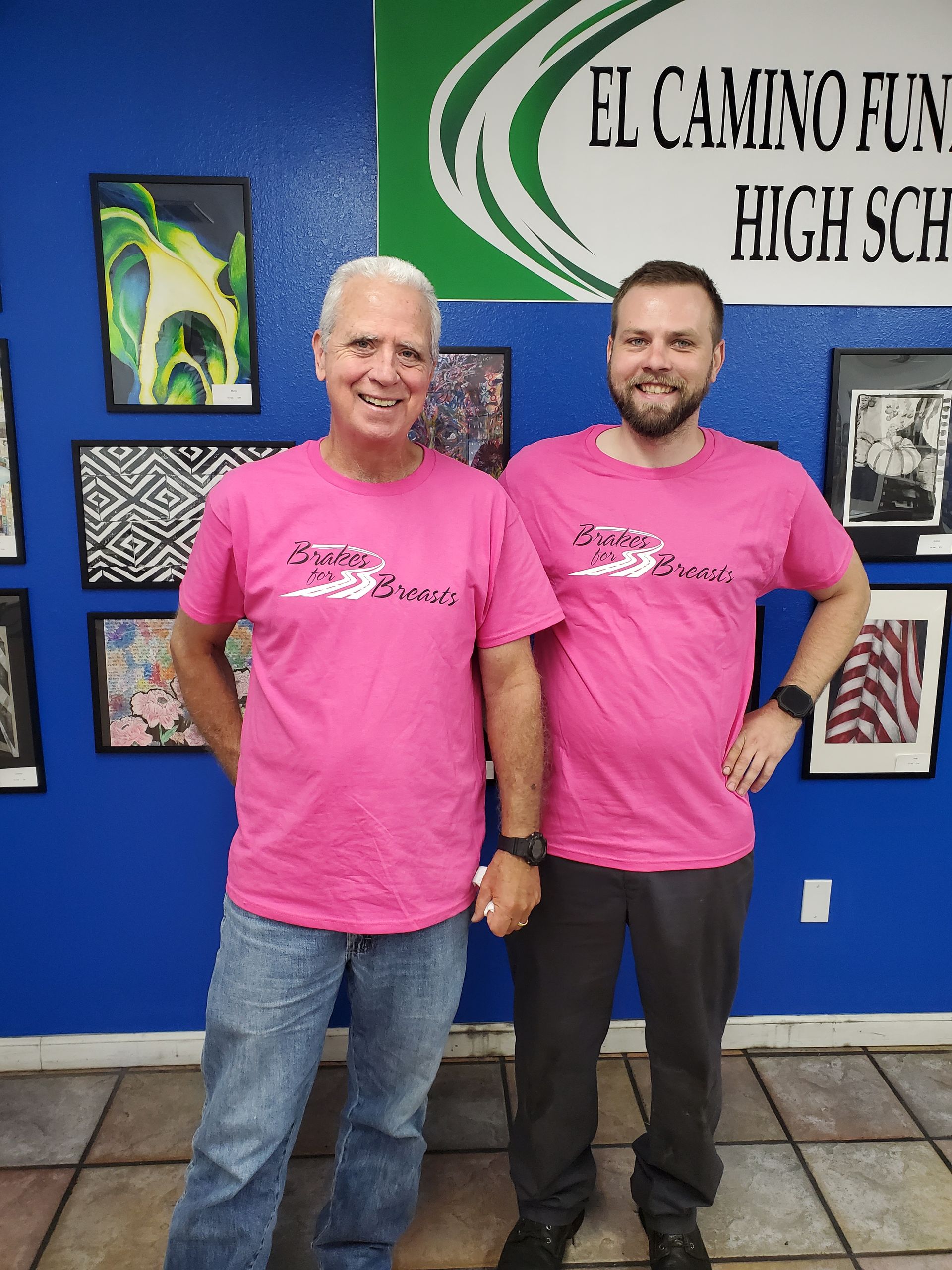Two men in pink shirts stand in front of a blue wall with framed art. One has grey hair and jeans, the other has a beard and dark pants. | Hal's Auto Care