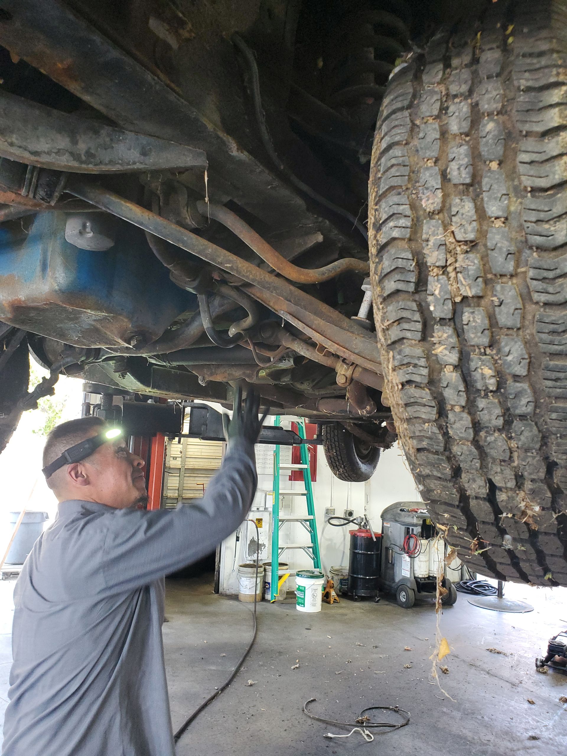 Mechanic under a vehicle, inspecting parts with a headlamp in a repair shop. | Hal's Auto Care
