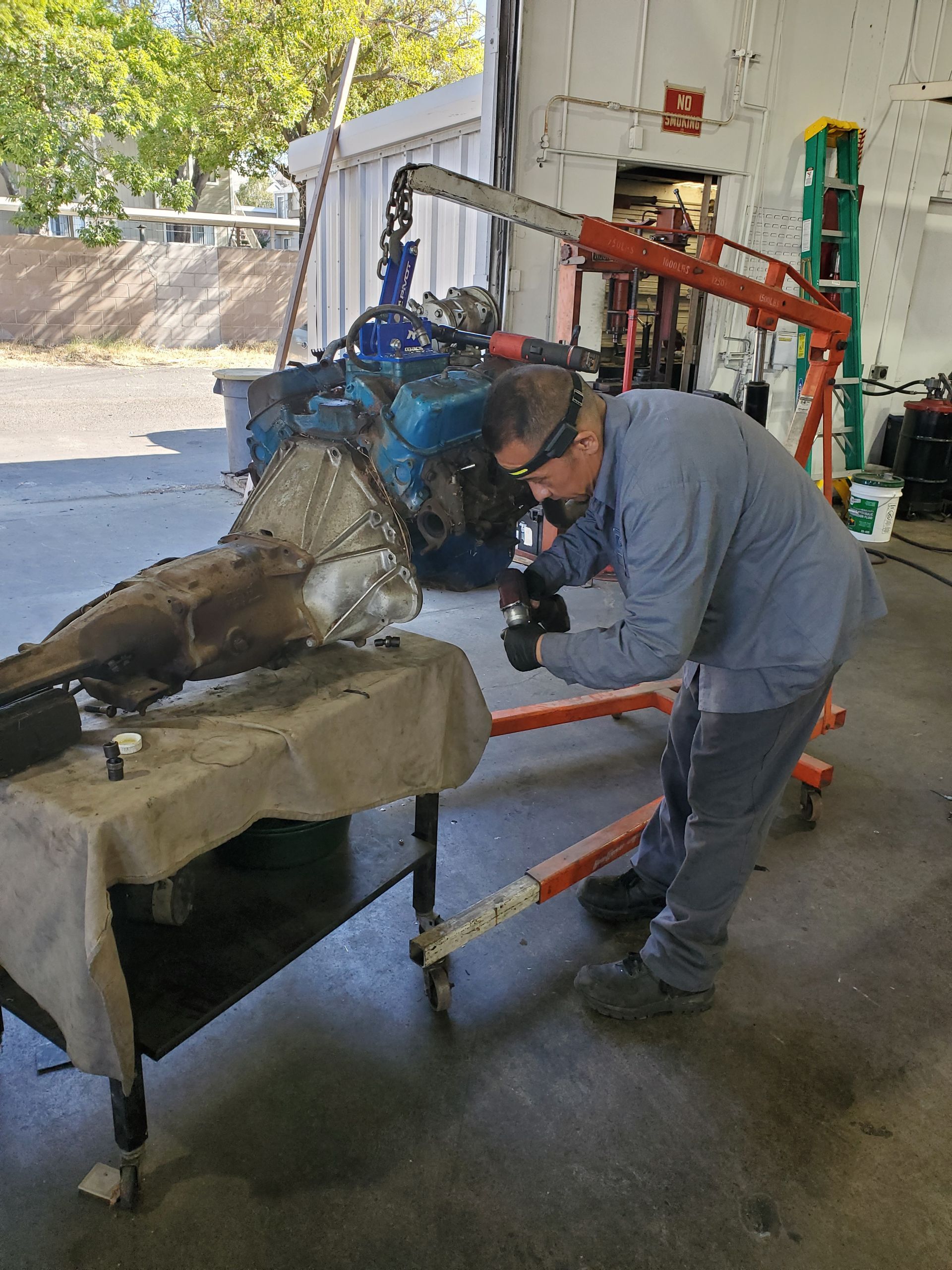 Mechanic working on an engine and transmission in a garage.  Engine hoist, table, and tools are present. | Hal's Auto Care