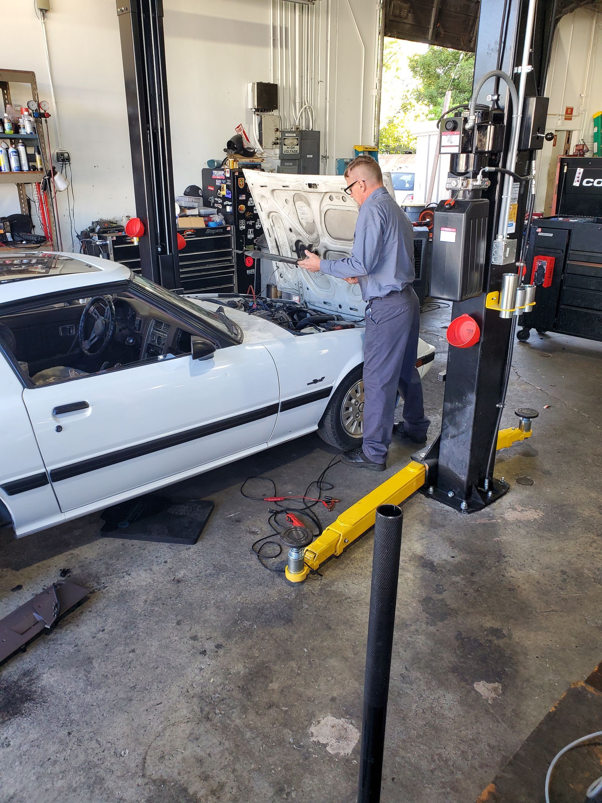Mechanic in gray uniform working on white car with open hood, in a garage. Car on a lift. | Hal's Auto Care