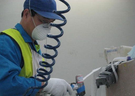 Factory worker wearing a mask, gloves, and blue uniform uses a tool on a white surface.