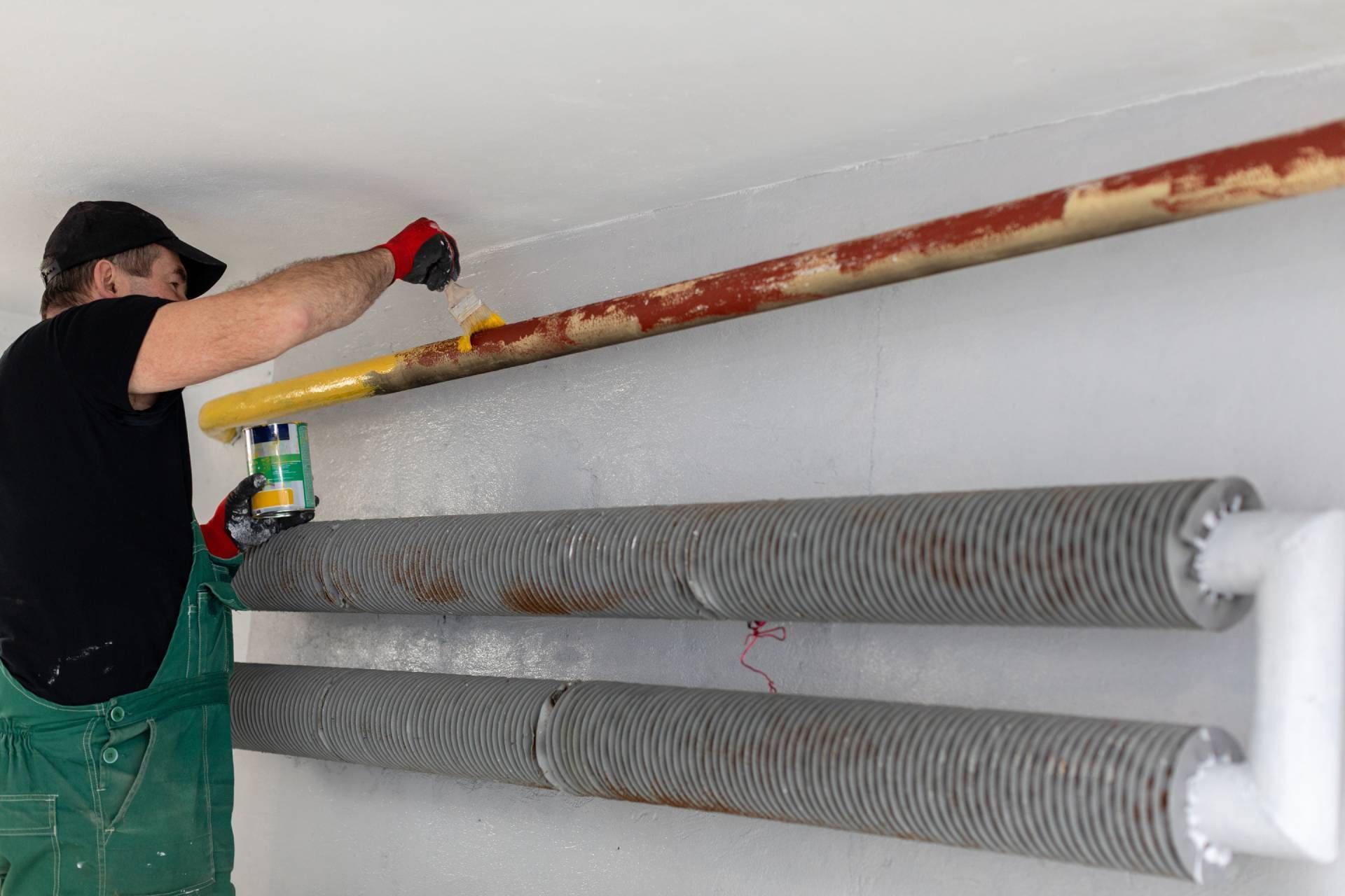 Man painting a brown metal tube on a white wall, standing near two gray, coiled tubes.