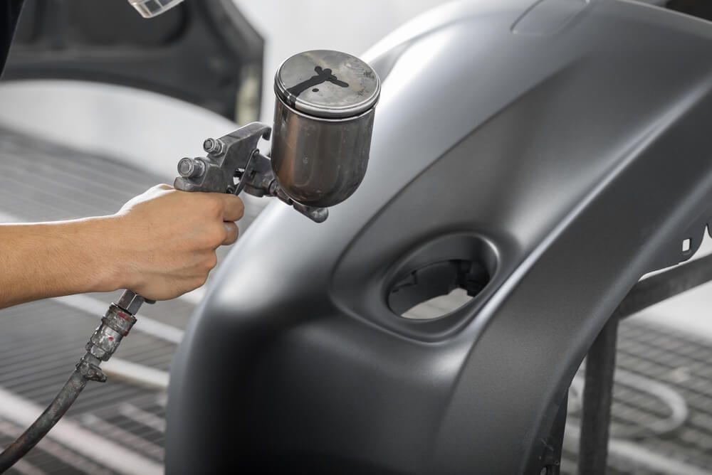 Person in protective suit spraying paint in a workshop; metal parts hanging.