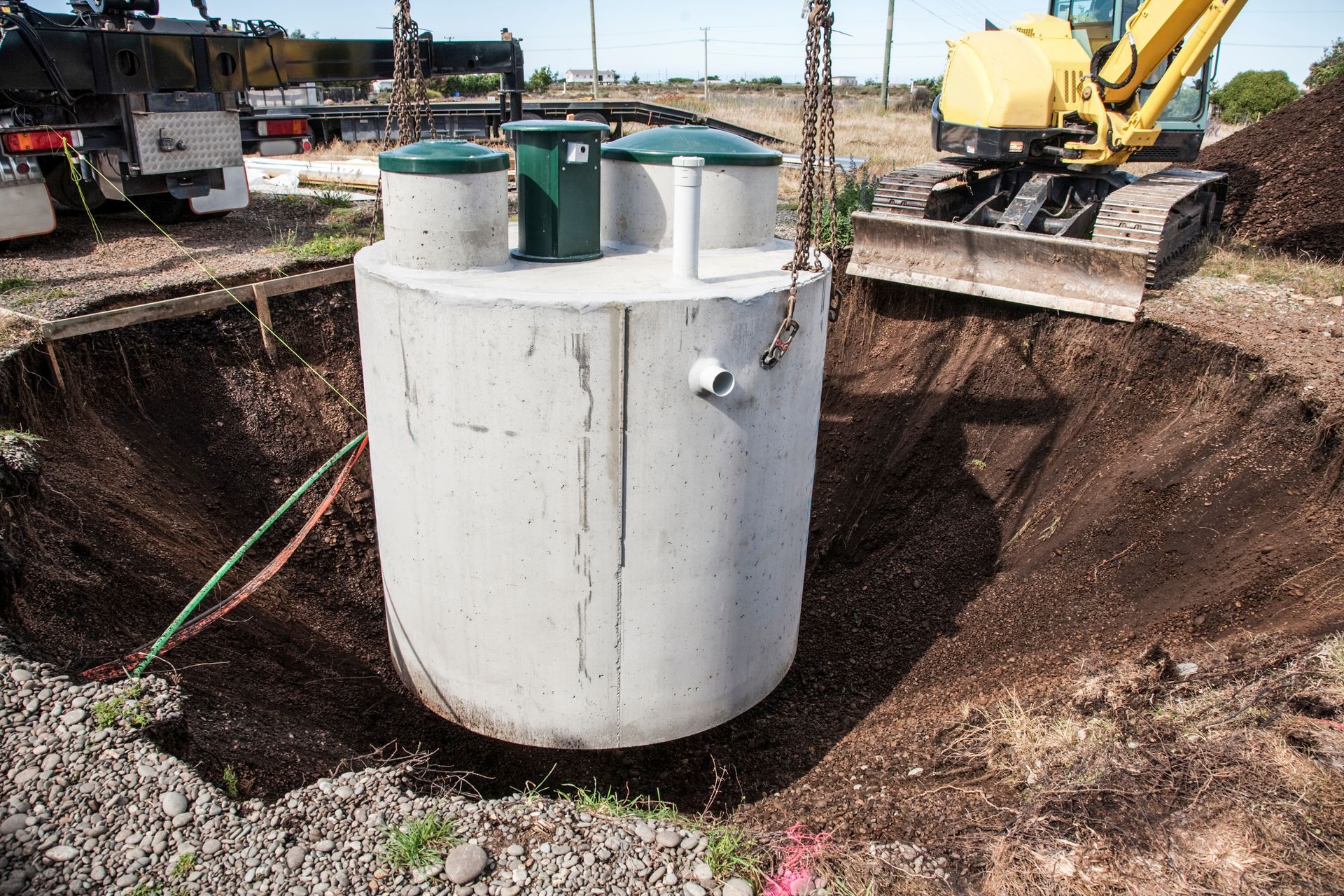 Concrete septic tank being lowered into a large excavation pit by crane; excavator and trucks in background.
