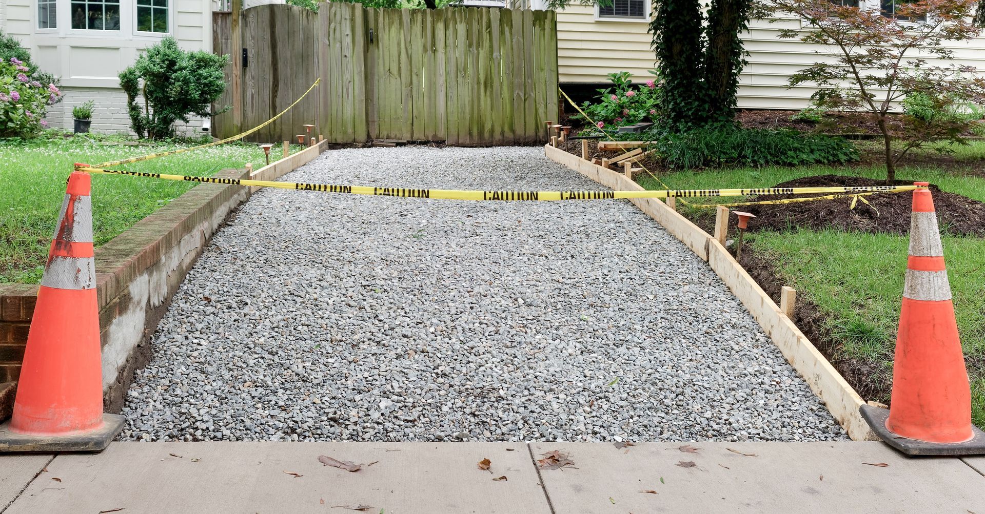 Gravel driveway under construction, bordered by wooden forms and caution tape, with orange cones at each end.