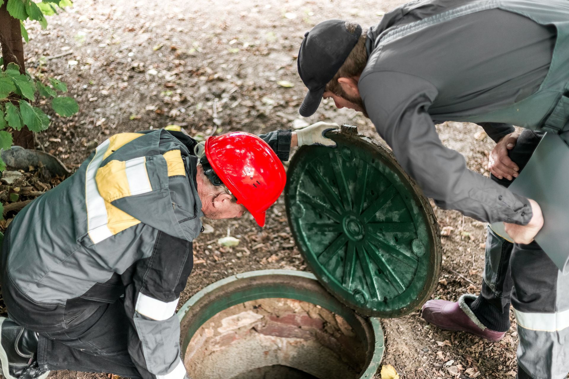 Two workers examine an open sewer manhole. One wears a red hard hat.