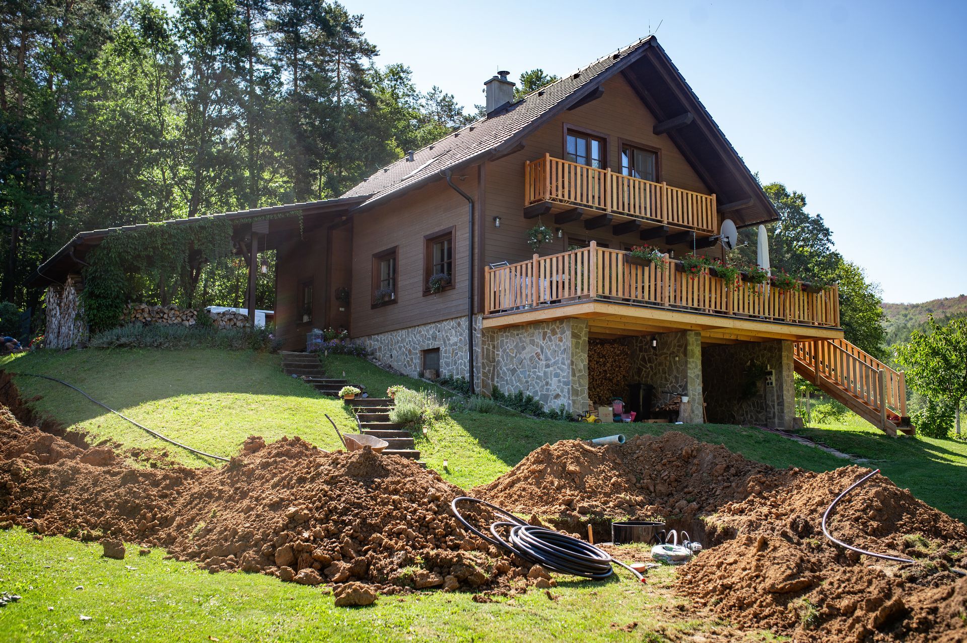 Brown house with stone base and wooden balconies, surrounded by piles of dirt and green grass.