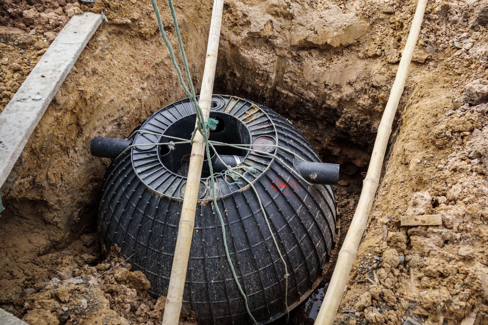 A large, black, ribbed plastic septic tank is partially installed in a dirt pit, with pipes attached to its sides.