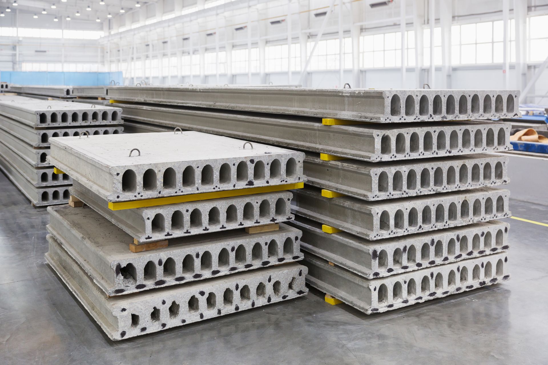 Stacks of gray concrete hollow-core slabs in a factory setting, ready for construction.