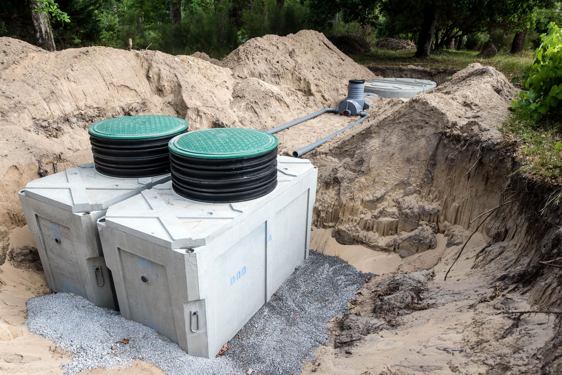 Two concrete septic tanks with green lids in an excavated area with sand, gravel, and connected piping.