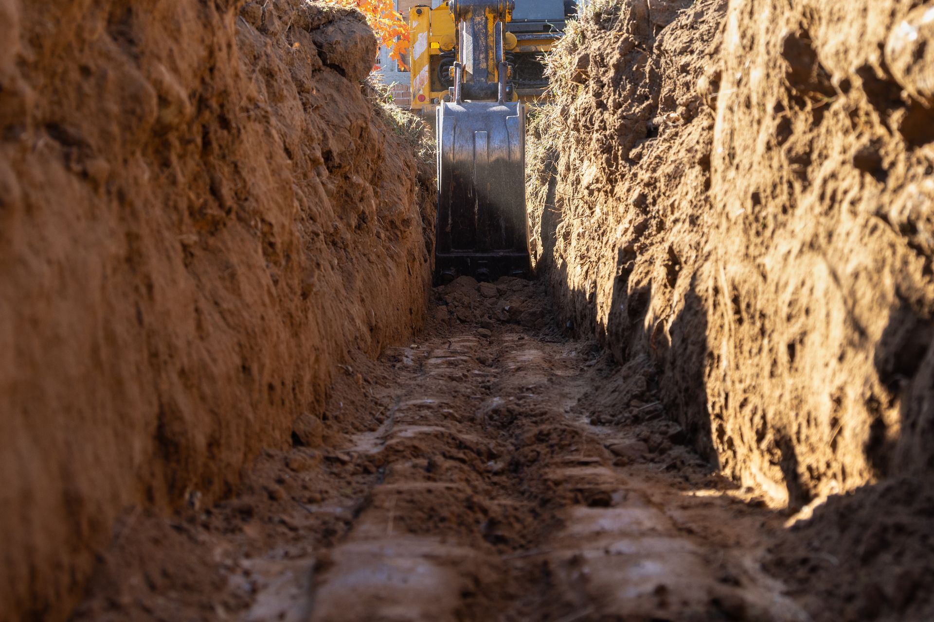 A deep trench being dug by a yellow excavator with its bucket lowered.