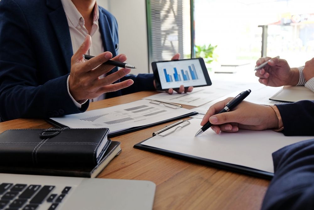 Reunião de negócios: gesticulando, revisando gráficos em tablet e papel, escrevendo em uma prancheta.