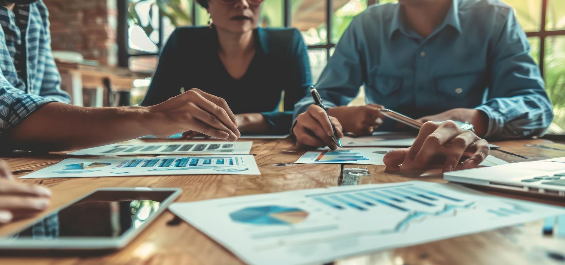 Pessoa apontando caneta para gráficos financeiros com outra pessoa em pé ao fundo.