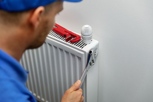 Man in blue shirt repairs a white radiator with a wrench.