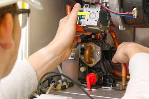 Technician repairs a gas boiler, focused on internal components, wearing glasses and a hard hat.
