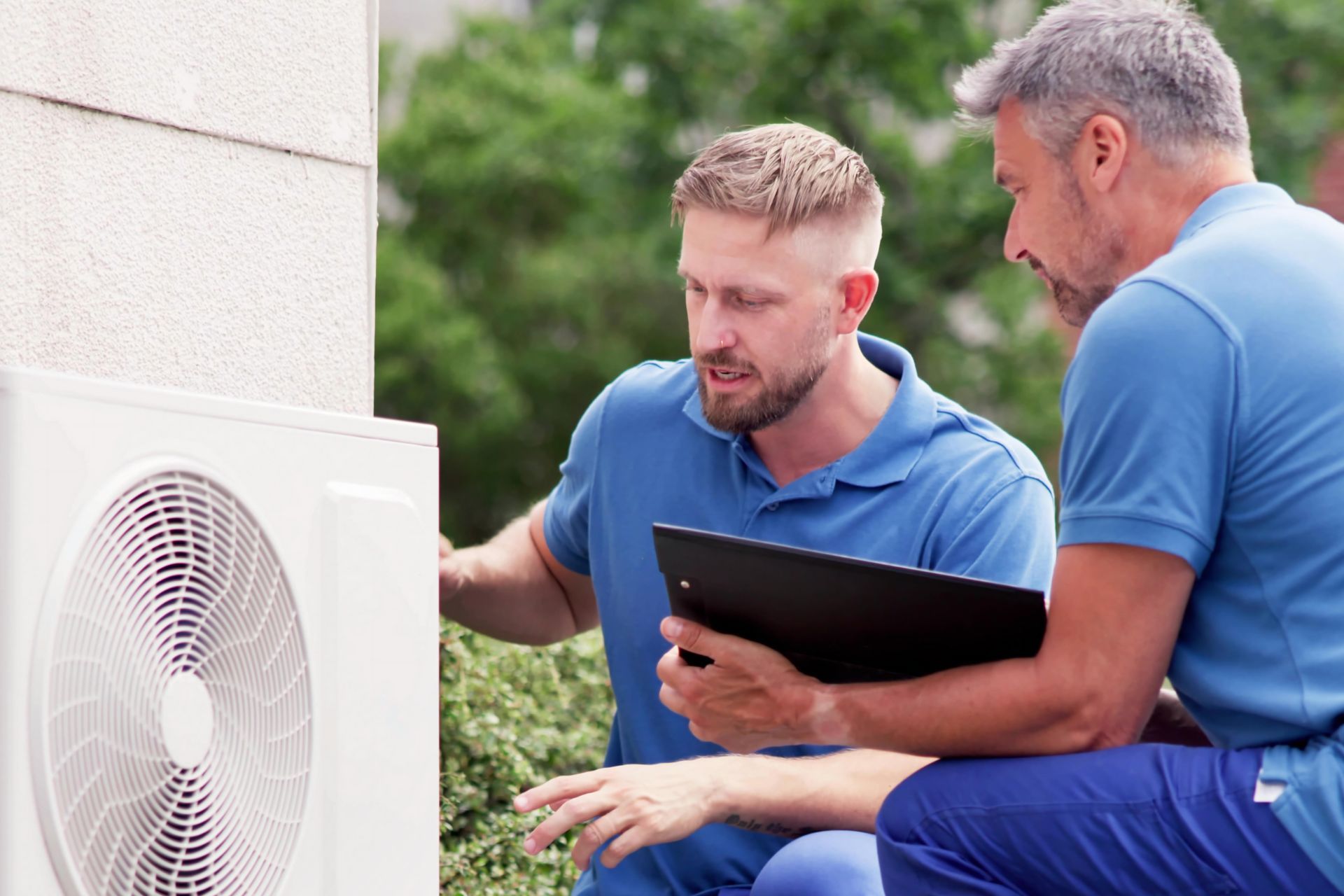 Two men are looking at a fan on the side of a building.