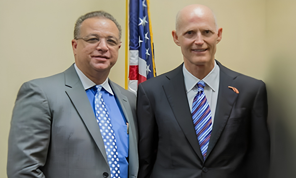 Two men in suits standing side by side in front of a U.S. flag and beige wall.