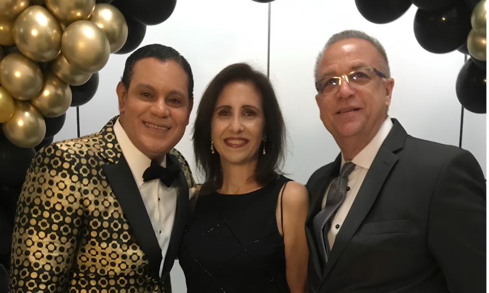 Three people smiling at a formal event, standing before black, gold, and silver balloon decorations.