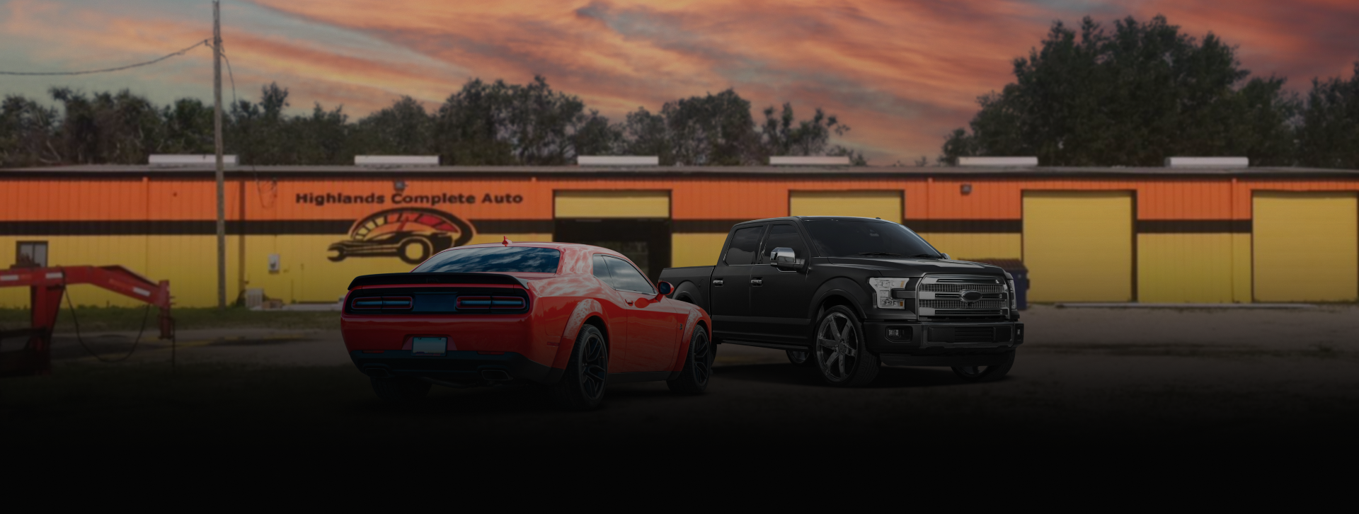 A red car and black truck parked outside a garage with yellow doors during sunset. | Highlands Complete Auto