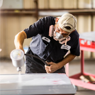 Man in protective gear sprays paint on a car part in a workshop. | Highlands Complete Auto