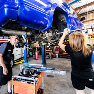 Two people working on a blue car in a garage. One person is using a wrench, the other is smiling. Tools on a cart. | Highlands Complete Auto