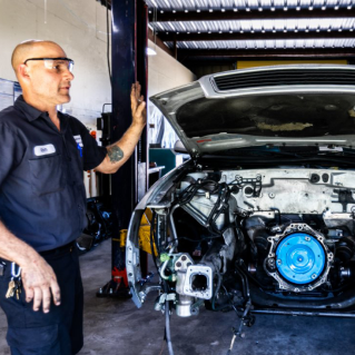 Mechanic gesturing at car engine bay in a shop. The car's front is disassembled. | Highlands Complete Auto
