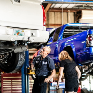 Mechanic and customer examine underside of lifted white truck in auto shop, blue truck in background. | Highlands Complete Auto