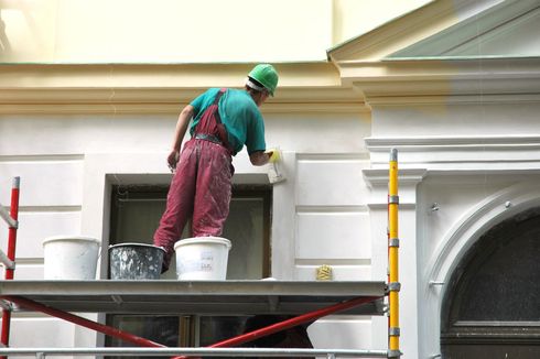 Imbianchino in tuta e casco, su un'impalcatura, mentre dipinge l'esterno di un edificio.
