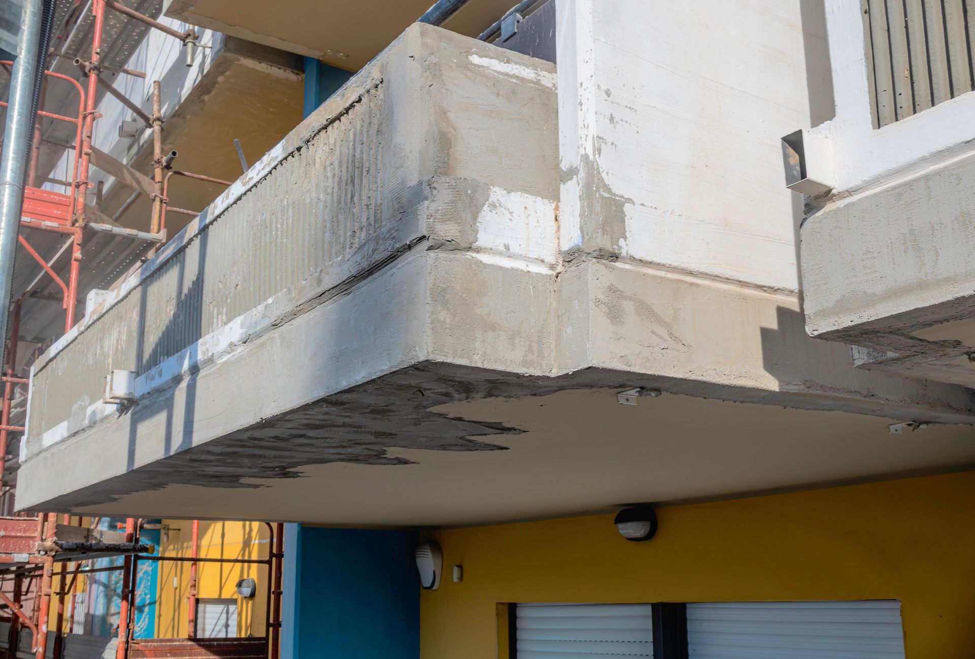 Balcone in cemento in fase di riparazione, con impalcature visibili e cemento fresco. L'edificio è dipinto di blu e giallo.