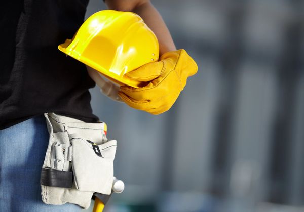 Operaio edile con in mano un casco giallo e un guanto; cintura portautensili visibile.
