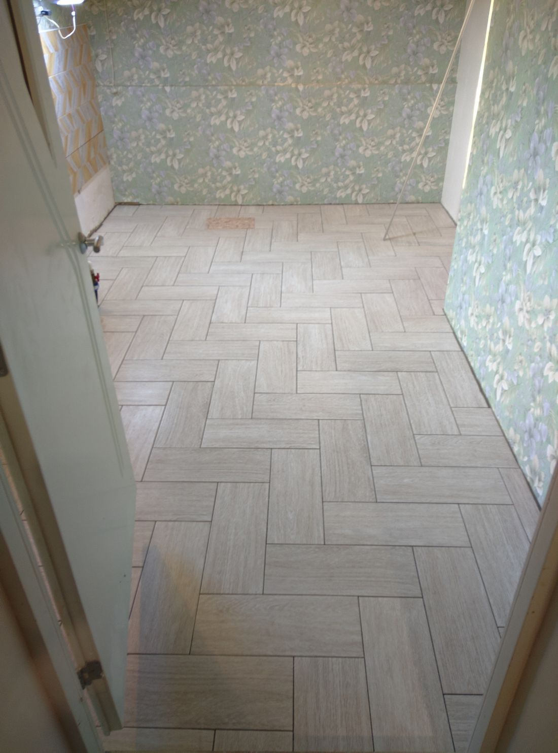 Camera con pavimento piastrellato a spina di pesce, carta da parati floreale e porta aperta.