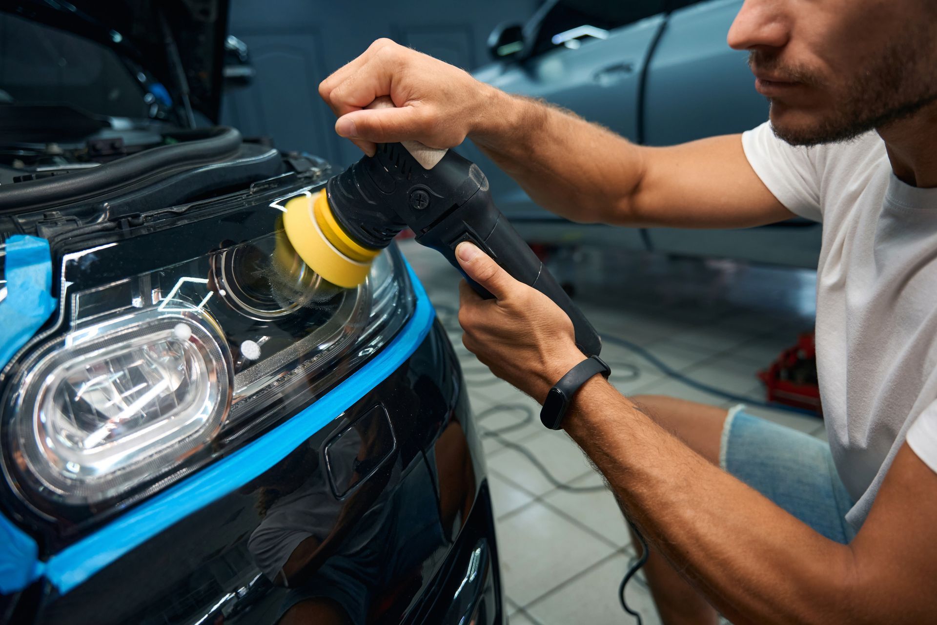 Person polishing a car headlight with a power tool; blue tape around edges.
