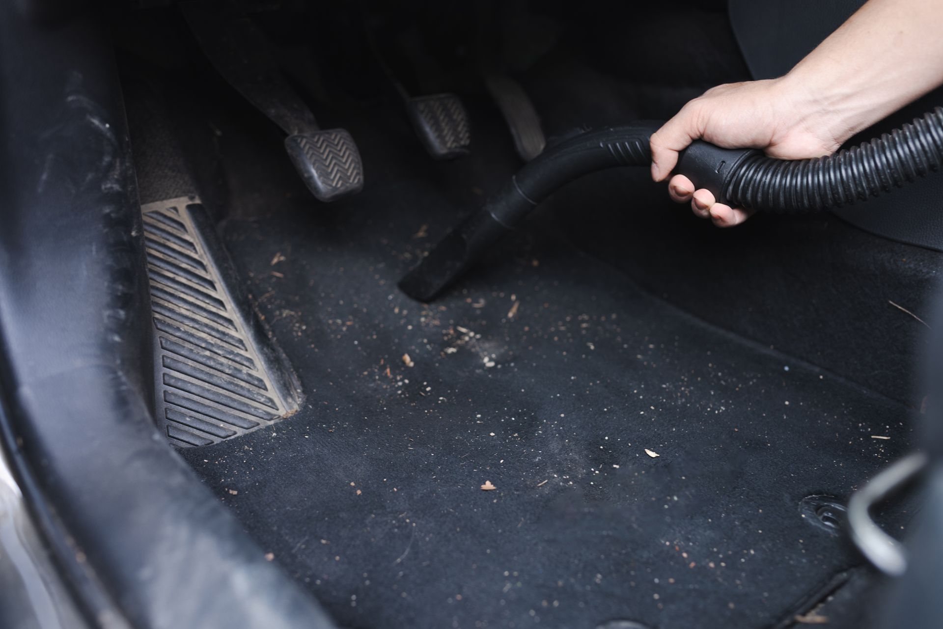 Hand vacuuming a car's black floor mat with debris, near foot pedals. Hand vacuuming a car's black floor mat with debris, near foot pedals.