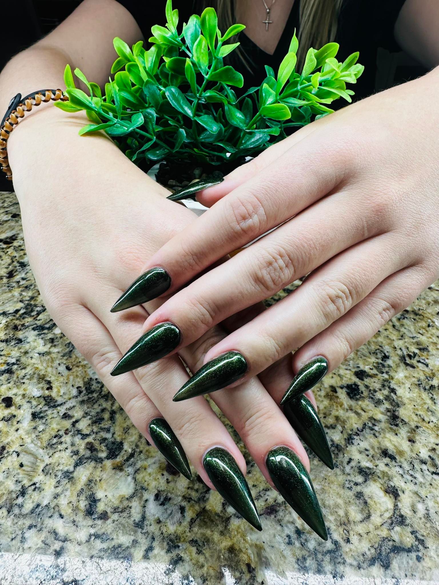 Hands with long dark green nails resting on a granite surface beside a small green plant