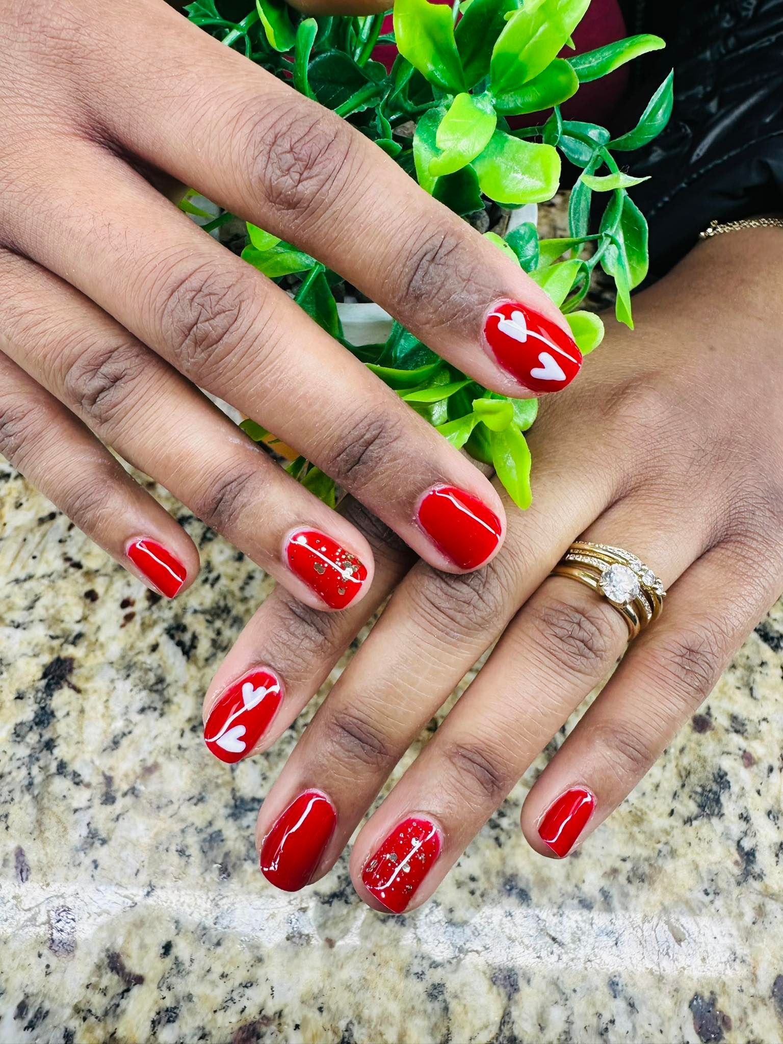 Hands with red nail art and white floral designs resting on a granite surface near green leaves.