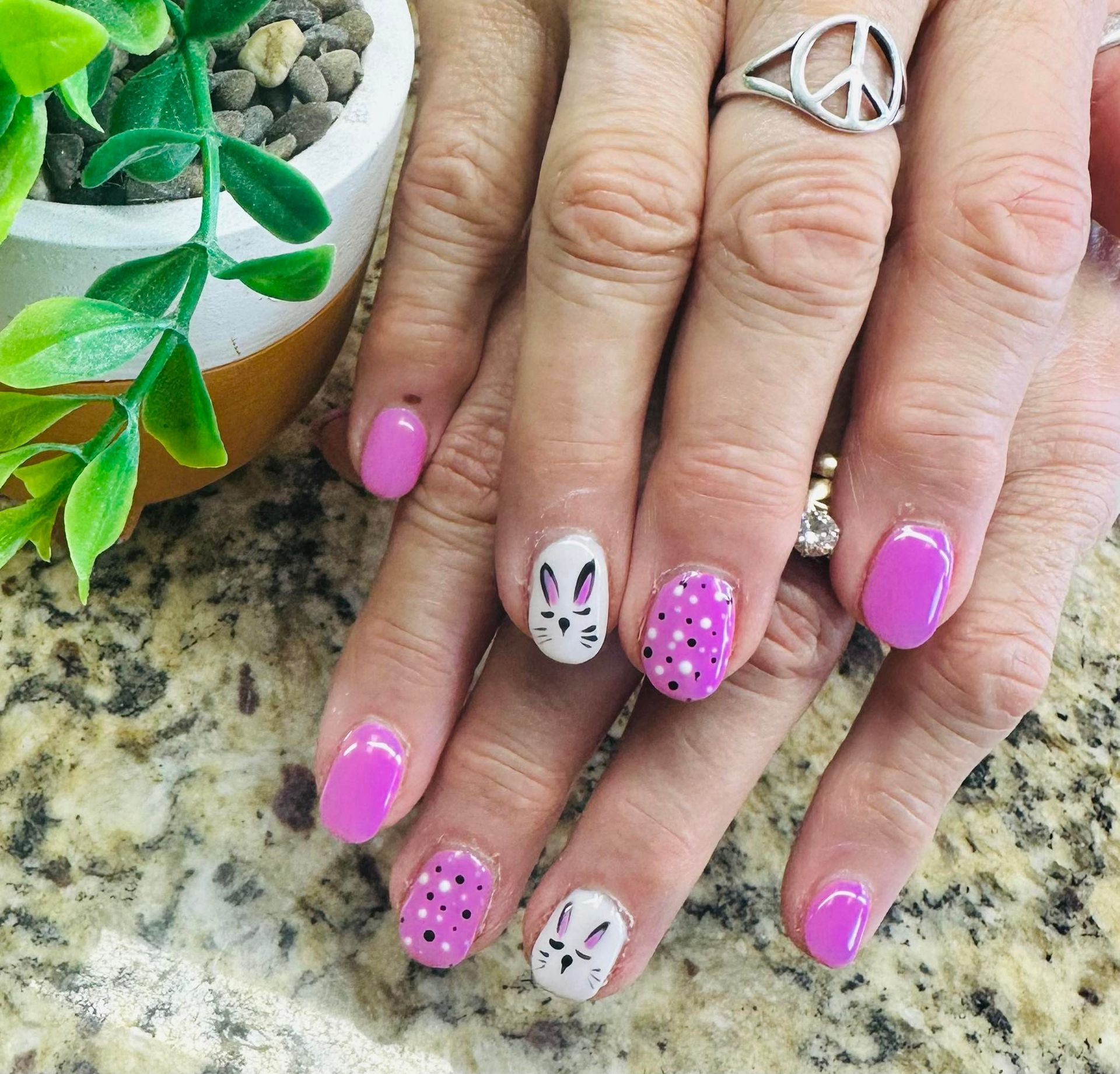 Hands with pink and white polka-dot nail art on a granite countertop, beside a small potted plant